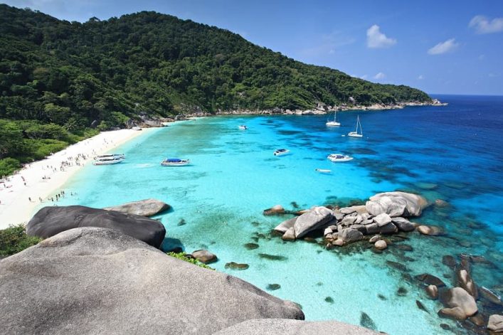 Strand mit weißem Sand, türkisblauem Wasser und felsiger Küste. Boote schwimmen im Wasser, dichte grüne Vegetation bedeckt den Hügel im Hintergrund. Felsen liegen im Vordergrund.