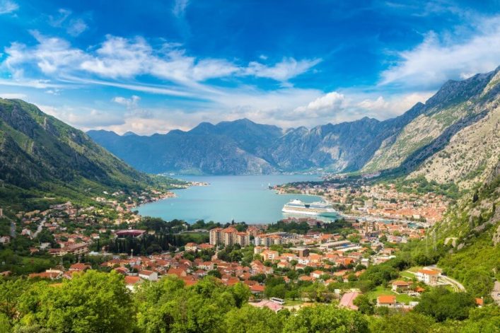 Blick auf die Bucht von Kotor, umgeben von grünen Bergen. Unten liegt eine Stadt mit roten Dächern und weißen Gebäuden. Der blaue Himmel ist von wenigen Wolken bedeckt. Meer erstreckt sich in der Mitte.