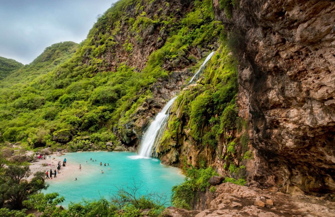 Ein Foto aus erhöhter Position von einem Wasserfall, der aus einem grünbewachsenen Berg entspringt und in dem türkisenen Wasser eines kleinen Sees mündet.