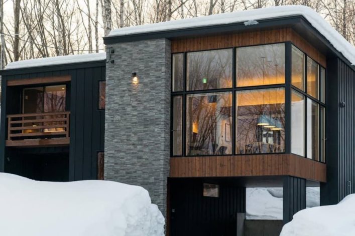Große, moderne Ferienvilla im japanischen Skigebiet Niseko. Die Fassade des stylischen Haus es besteht aus grauem Stein, Holzpanelen und einer riesigen Fensterfront. Im Vordergrund ist eine sehr hohe Schneedecke zu sehen.