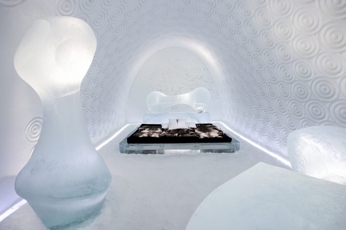 Ein schneebedecktes Zimmer im Eishotel mit einem großen Eisskulpturenpaar links und rechts, einem Bett in der Mitte und einer strukturierten Wand mit kreisförmigen Mustern im Hintergrund.