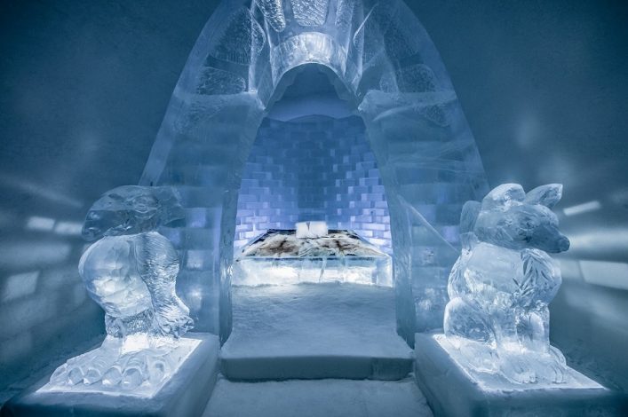 In einem Eishotel steht ein Bett aus Eis in der Mitte, umrahmt von kunstvollen Eisskulpturen links und rechts. Der Raum wird durch einen Eisbogen im Hintergrund hervorgehoben.