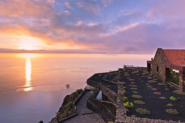Ein Sonnenuntergang über dem Meer an einer mit Pflanzen bewachsenen Terrassenklippe auf El Hierro, einem beliebten Ziel auf der Kreuzfahrtroute der Kanarischen Inseln. Links liegt das Meer, rechts ein Steinhaus mit roten Ziegeln.