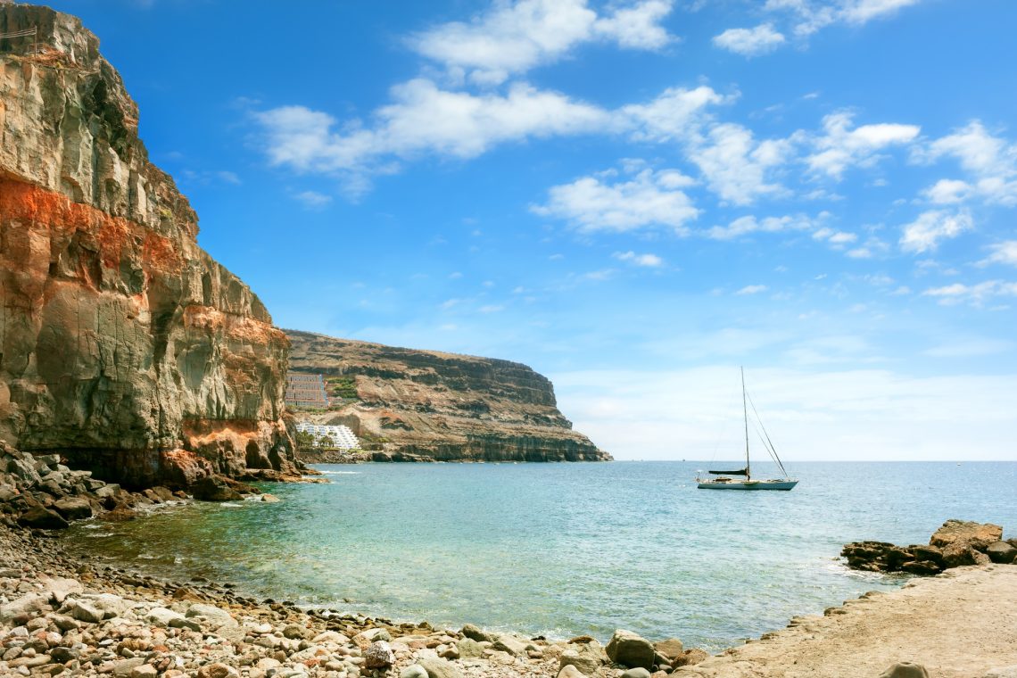 Foto einer Bucht auf Gran Canaria. Am unteren Teil des Bildschirms ist Sandstrand, dann beginnt das Meer. Links sind Klippen zu sehen. Auf dem Wasser liegt ein Segelboot vor Anker.