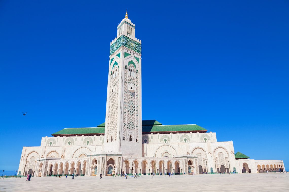 Die Hassan-II.-Moschee in Casablanca mit ihrem hohen, reich verzierten Minarett erhebt sich vor strahlend blauem Himmel. Die Moschee ist umgeben von einem großen Platz mit wenigen Menschen.