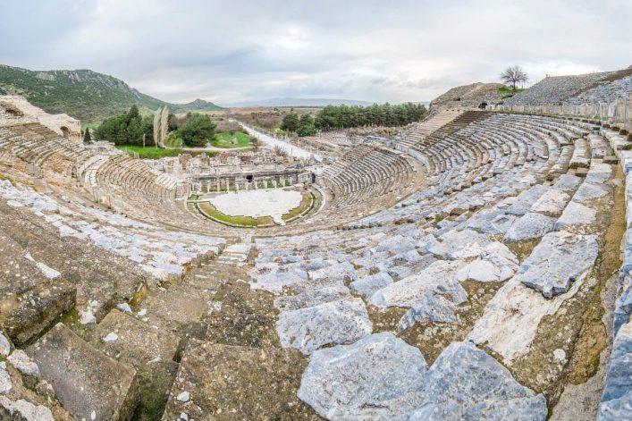 Antikes Steintheater in Ephesos in der Türkischen Ägäis mit abgestuften Sitzreihen, die sich bogenförmig um eine zentrale Bühne legen, umgeben von Hügeln.