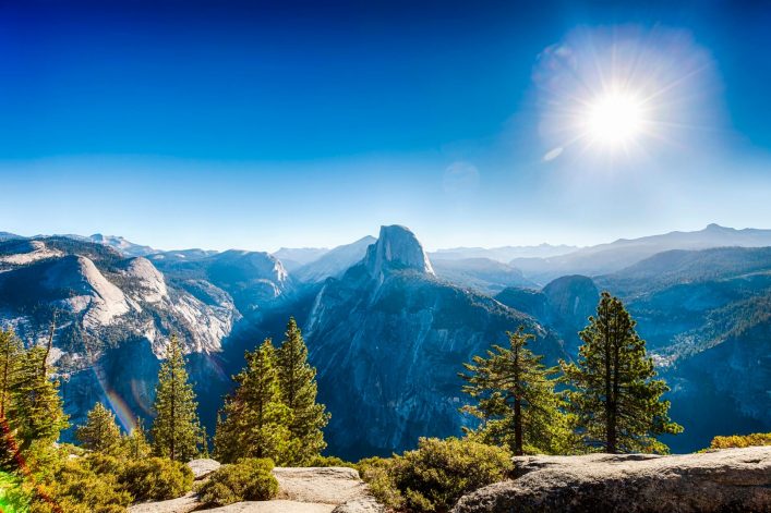 Blick vom Glacier Point auf das Yosemite-Tal mit dem markanten Half Dome im Zentrum, umgeben von bewaldeten Bergen. Im Vordergrund grüne Bäume und Sträucher, oben strahlende Sonne am klaren Himmel.