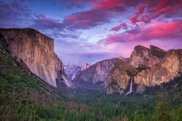 Blick auf das Yosemite-Tal: links markante Felswand El Capitan, mittig ferne Berge, rechts Wasserfall vor felsiger Kulisse. Der Himmel ist bunt, überwiegend lila und rosa, unten dichter Wald.