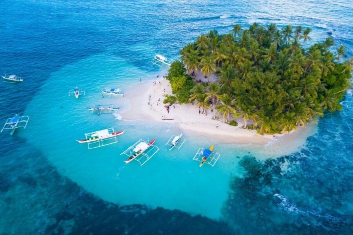 Eine kleine, palmengesäumte Insel mit weißem Sand in türkisfarbenem Wasser. Mehrere bunte Boote sind um die Insel verteilt. Menschen spazieren am Strand unter Palmen, leicht bewölkter Himmel.