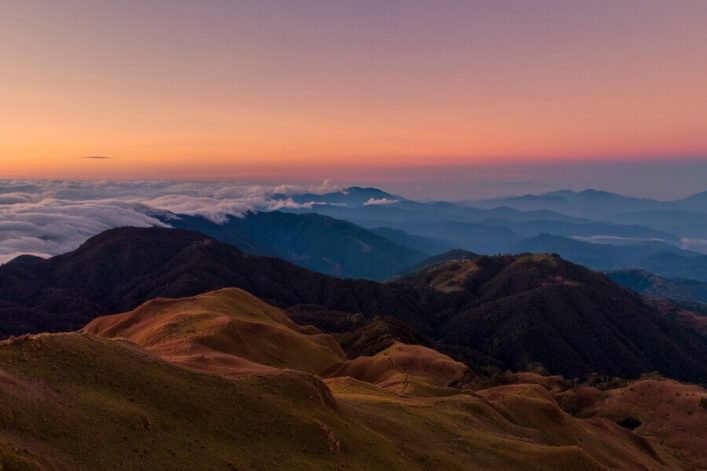 Berge mit sanften Hügeln im Vordergrund und nebligen Wolken im mittleren Bereich. Der Himmel ist von sanftem Orange bei Sonnenaufgang beleuchtet, und ferne Bergketten verblassen in Blau.