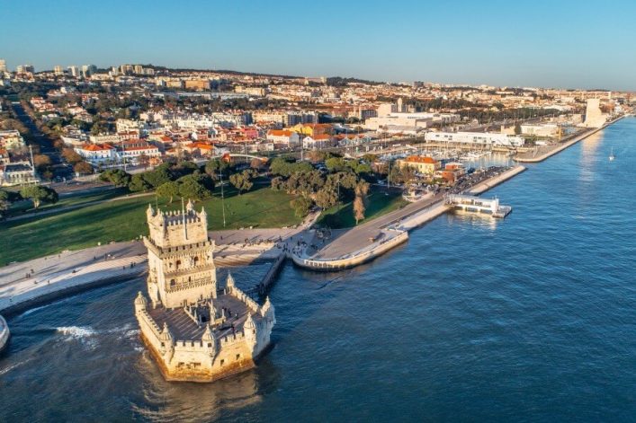 Luftaufnahme der Küste von Lissabon mit dem berühmten Belém-Turm im Vordergrund, umgeben von Wasser. Der gepflegte Uferbereich zieht sich in Richtung der Stadt, die als beliebtes Kreuzfahrtziel bekannt ist.