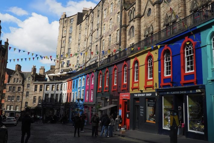 Bunte Häuserfronten in kräftigen Farben wie Blau, Rosa und Gelb reihen sich entlang der Victoria Street in Edinburgh. Links erheben sich historische Gebäude. Eine bunte Girlande verbindet die Gebäude.