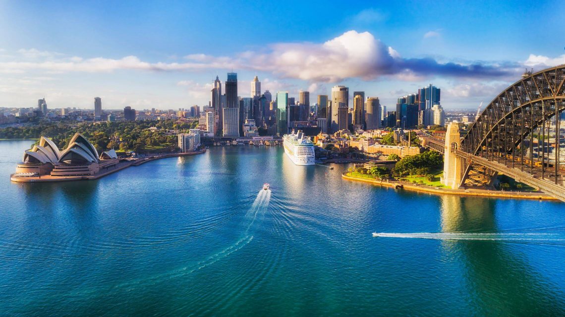 Blick auf den Hafen von Sydney mit Opernhaus links und Harbour Bridge rechts. Im Zentrum der Skyline liegt ein großes Kreuzfahrtschiff, ideal für eine Dezember-Kreuzfahrt unter blauem Himmel und wenigen Wolken.