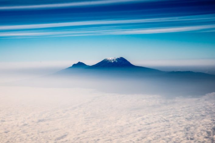 Freistehender schneebedeckter Berg Kilimandscharo in der Bildmitte, unter einem klaren blauen Himmel mit vereinzelten Wolkenstreifen. Dichter Wolkenteppich bedeckt den unteren Bildbereich.