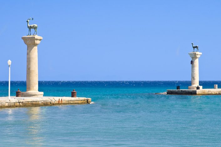 Zwei Säulen stehen auf Molen am Meer, jede mit einer Hirschstatue gekrönt. Klare blaue See im Vordergrund, dahinter leuchtet der Himmel. Links eine Straßenlaterne, beides bei Rhodos.