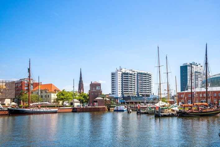 Am Ufer des Hafens in Bremerhaven liegen mehrere Segelschiffe vertäut. Im Hintergrund erheben sich moderne Gebäude und ein Kirchturm. Diese Stadt gehört zu den beliebtesten Kreuzfahrtzielen Nordeuropas.