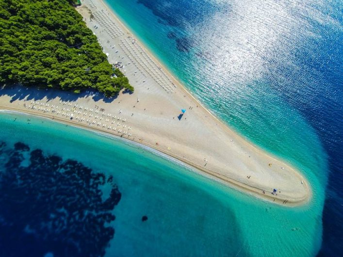 Luftaufnahme von Zlatni Rat auf Brač: schmale Kieslandzunge im Meer, Pinienwald im Hintergrund, türkis bis dunkelblaues Wasser.