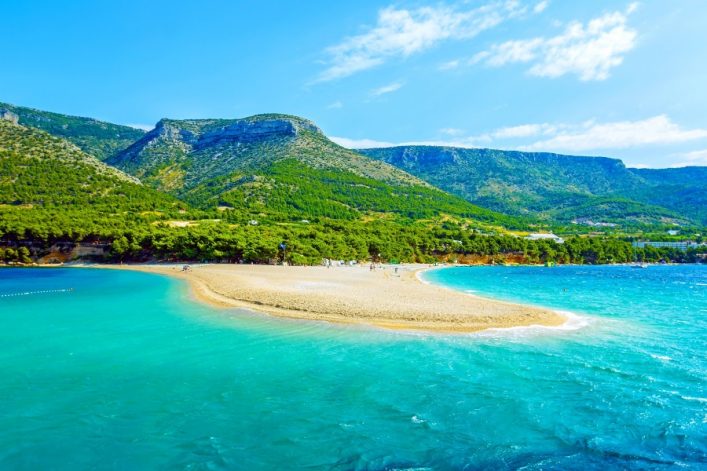Zlatni Rat auf Brač vom Wasser aus: sandig‑kiesige Landzunge im türkisblauen Meer, dahinter grünes Gebirge.