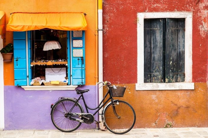 Buntes Fahrrad lehnt an farbenfroher Hauswand auf Burano. Links hellblaues Fenster mit orangefarbenem Vordach, rechts dunkles Fenster vor rotbrauner Wand. Pflastersteine bilden den Boden.