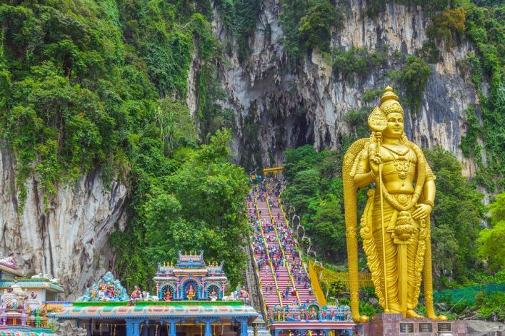 Goldene Statue von Lord Murugan rechts, bunte Treppe in der Mitte führt zu Höhle, umgeben von grünen Bäumen und Felsen. Vordergrund zeigt kunstvoll verziertes Tempeldach.