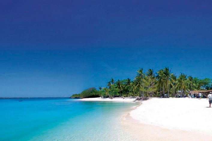 Strahlend blauer Himmel über türkisblauem Meer. Rechts ein weißer Sandstrand mit Palmen am Rand. Eine Person spaziert am Wasser entlang. Im Hintergrund Hütten unter den Palmen.