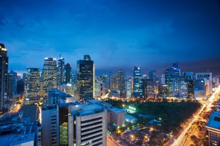 Hochhäuser der Makati-Skyline bei Dämmerung, verteilt im Bildzentrum. Unten ein Park und Straßen mit Lichtern, die nach rechts führen. Der Himmel ist dunkelblau und klar.