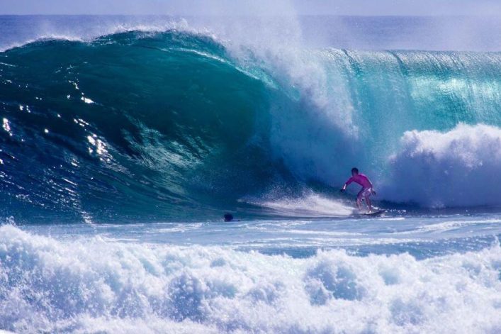 Surfer in pinkem Outfit reitet auf einer mächtigen türkis-blauen Welle; die Welle türmt sich links groß auf, während der Schaum unten rechts das Wasser aufwirbelt.