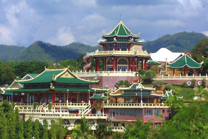 Prächtiger taoistischer Tempel mit grünen Dächern und roten Säulen, umgeben von üppigem Grün; im Hintergrund sanfte Hügel unter einem bewölkten Himmel. Farbdetaillierte Architektur in der Natur.