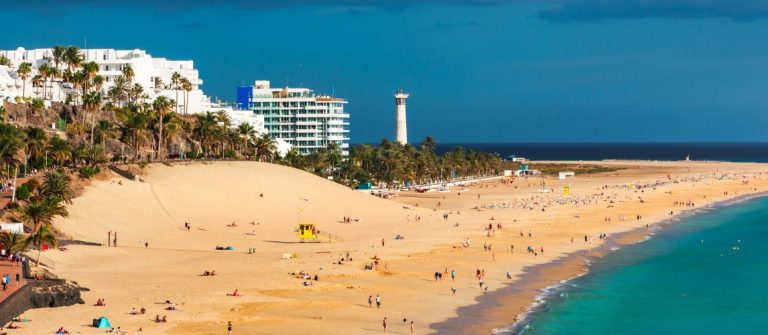 Strand von Morro del Jable auf Fuerteventura mit goldenem Sand und türkisblauem Meer. Im Hintergrund stehen weiße Gebäude und ein Leuchtturm, Palmen sind auf der linken Bildseite zu sehen.