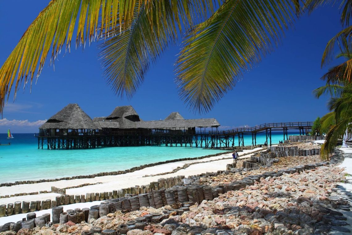 Ein Steg mit strohgedeckten Hütten erstreckt sich ins türkisblaue Meer. Im Vordergrund sind ein weißer Sandstrand, Felsen und Palmenblätter. Rechts geht ein Mensch spazieren, links gleitet ein Segelboot.