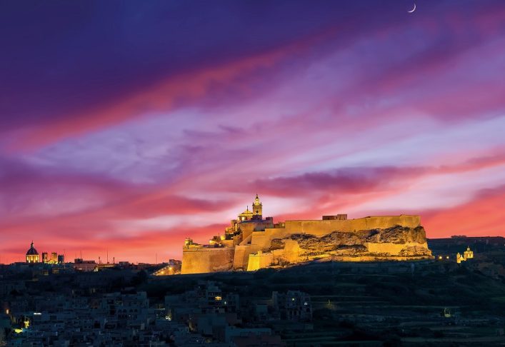 Im Vordergrund leuchtet eine beleuchtete Festung auf einem Hügel, im Hintergrund ein farbenfroher Sonnenuntergang mit violetten und roten Wolken, rechts oben ein schmaler Halbmond am Himmel.