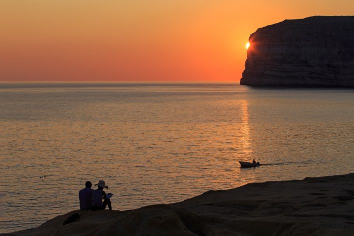 Sonnenuntergang über dem Meer. Rechts eine Felsklippe mit der Sonne, die dahinter verschwindet. Links vorne sitzen zwei Personen auf einem Felsen. Ein kleines Boot fährt auf dem Wasser.