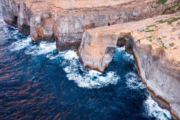 Im Vordergrund ragt ein beeindruckendes Felsentor ins blaue Meer, Wellen brechen daran. Der Hintergrund zeigt eine zerklüftete Steilküste, die von oben fotografiert wurde.