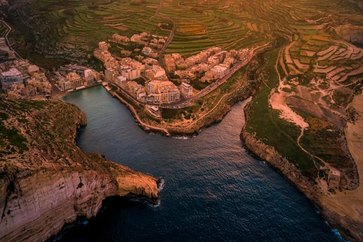 Luftaufnahme einer Küstenstadt mit Gebäuden an einer Bucht, umgeben von grünen Terrassenfeldern. Der Ozean erstreckt sich im Vordergrund, während Hügel das Hinterland prägen. Sonnenlicht wirft warme Töne.