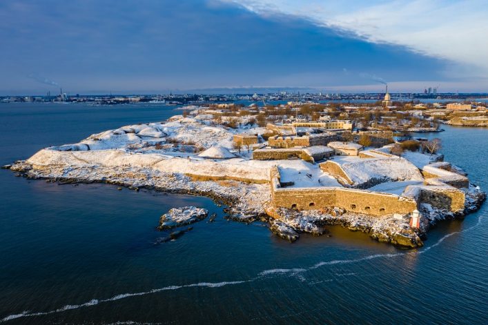 Luftaufnahme von Suomenlinna im Winter. Die schneebedeckte Festung liegt auf einer Insel, umgeben von blauem Meer und im Hintergrund ist die Stadt Helsinki mit Gebäuden zu sehen.