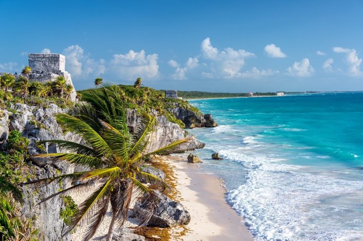 Strand von Tulum in Mexiko. Sanfte Wellen des türkisfarbenen Meers schwappen an einen hellen, weichen Strand. Auf Felsklippen oberhalb des Strandes wachsen Palmen, außerdem sind antike, weiße Ruinen zu sehen.