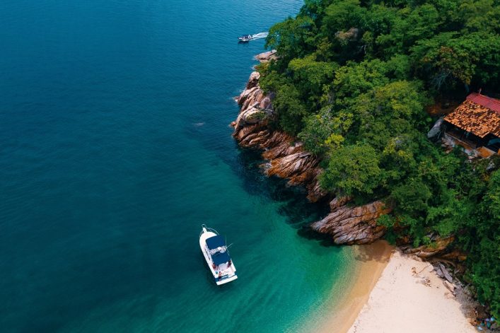 Eine Drohnenaufnahme zeigt einen weißen Katamaran in türkisblauem Wasser vor einem bewaldeten Hügel. Am Hang ragt ein Haus mit rotem Dach heraus. Ideal für eine April-Kreuzfahrt nach Majahuitas.