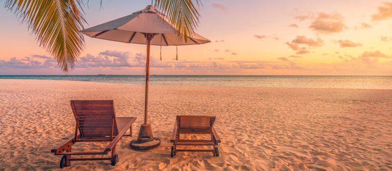 Zwei leere Holzliegen und ein Sonnenschirm stehen auf hellem Sandstrand, eingerahmt von Palmen, mit Blick auf das ruhige Meer und einen orangefarbenen Himmel bei Sonnenuntergang.
