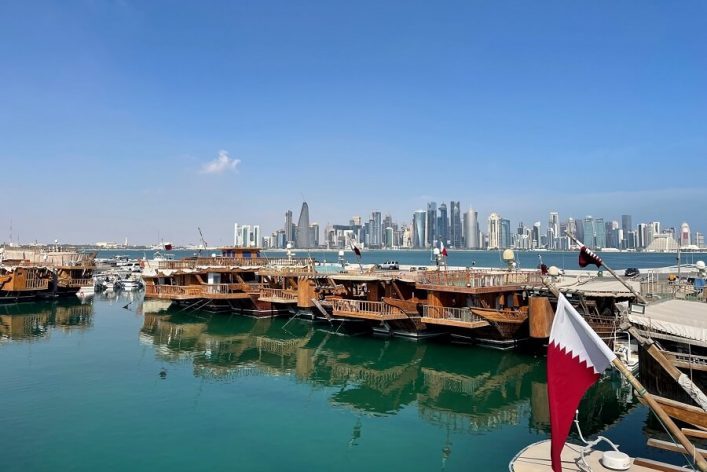 Traditionelle Holzboote schwimmen im klaren Wasser des Dhow-Hafens in Katar. Im Hintergrund erhebt sich die beeindruckende Skyline von Doha. Eine katarische Flagge weht im Vordergrund.