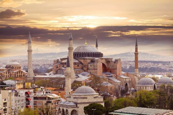 Eine große historische Kuppelmoschee mit vier Minaretten steht inmitten dichter Stadtbebauung, im Hintergrund Hügel mit Gebäuden und ein wolkiger Himmel bei Sonnenuntergang.