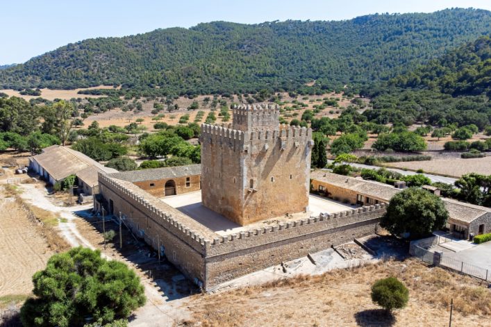 Ein quadratischer Steinturm mit Zinnen steht zentral, umgeben von einer rechteckigen Mauer. Im Hintergrund erstreckt sich eine bewaldete Hügellandschaft, umgeben von Feldern und vereinzelt stehenden Bäumen.