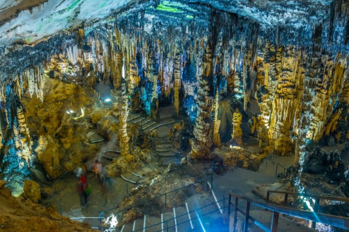 In der Tropfsteinhöhle ragen beeindruckende Stalagmiten und Stalaktiten von Boden und Decke. Ein beleuchteter Pfad führt durch die Höhle, wo Menschen die faszinierende Natur betrachten.