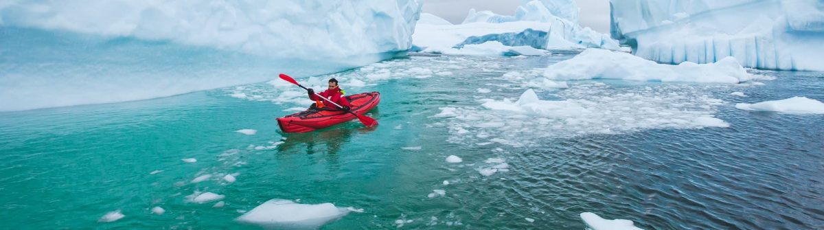 Ein Kayak Fahrer paddelt in einem roten Kayak entlang von Eisblöcken durch das Meer der Antarktis