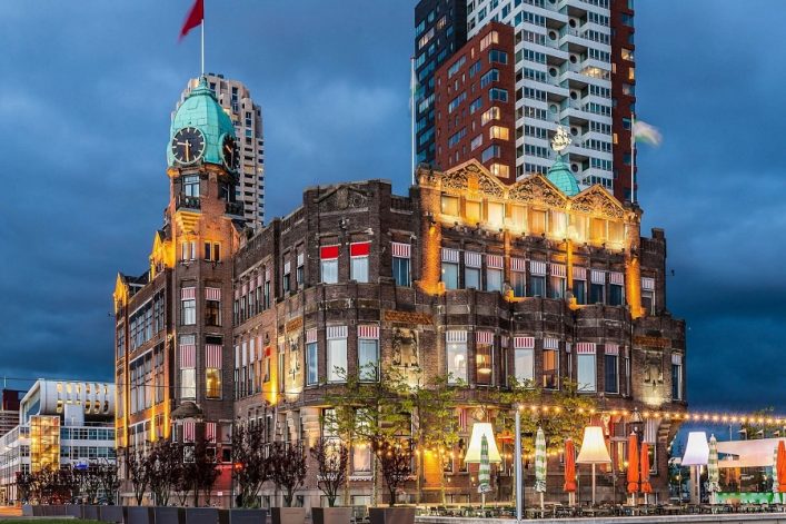 Historisches Gebäude mit Uhrturm und grüner Kuppel vor modernem Hochhaus bei Abenddämmerung. Das Gebäude ist beleuchtet, im Vordergrund sind Bäume und Lampen zu sehen.