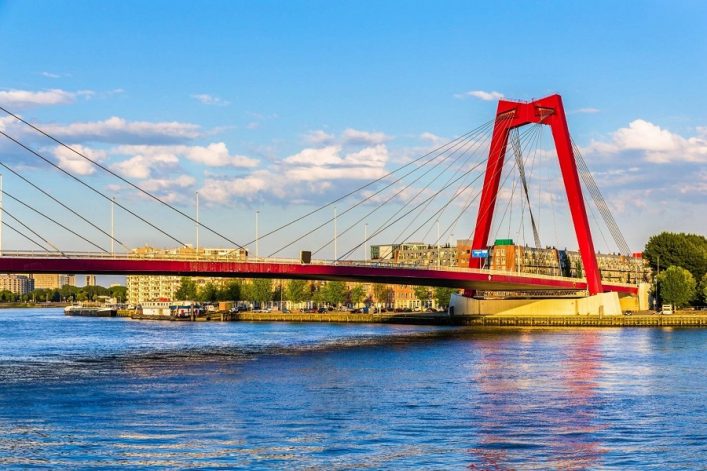 Rote Hängebrücke mit markanten Balken und Streben über einem Fluss; im Hintergrund moderne Gebäude und blauer Himmel mit weißen Wolken; Wasser spiegelt den Himmel.