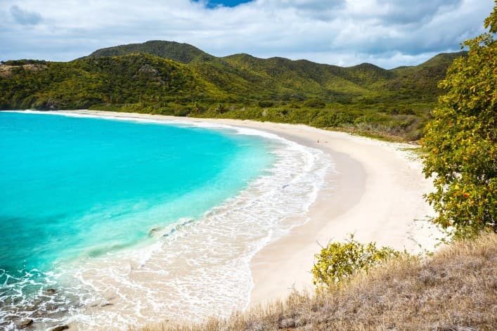 Hier wurde der Rendezvous Strand auf Antigua aus der Höhe fotografiert, sodass man den Strand, der sich sichelförmig um das türkisfarbene Meerwasser schmiegt, erkennt. Im Hintergrund ist die grüne Felslandschaft zu erkennen.