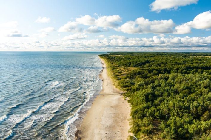 Blick von oben auf die Küste der Ostsee. Rechts sieht man dichte grüne Wälder, mittig läuft ein weißer Sandstrand durch das Bild und auf der linken Hälfte ist das Meer zu sehen. Die Wellen brechen vor der Küste. Der Himmel ist blau und von einigen Wolken durchzogen.