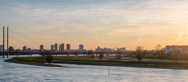 Panorama Aufnahme der Düsseldorfer Skyline bei Sonnenuntergang. Der gesamte Vordergrund des Bildes wird vom Rhein eingenommen, im Hintergrund reihen sich zahlreiche Gebäude aneinander. Besonders prominent sind der Rheinturm und die sich anschließende Rheinkniebrücke.