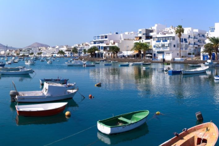 Kleine Boote schwimmen auf blauem Wasser in einem Hafen; im Hintergrund weiße Gebäude und Palmen. Beliebte Kreuzfahrtrouten führen zu solchen malerischen Orten mit typischen Küstenstädten.