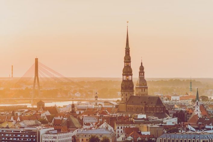 Die Petrikirche in Riga mit ihrem 132 Meter hohen Turm überragt die Altstadt. Im Hintergrund ist die Vanšu-Brücke und ein sanft leuchtender Sonnenuntergang zu sehen. Beliebte Kreuzfahrtrouten führen oft hierher.